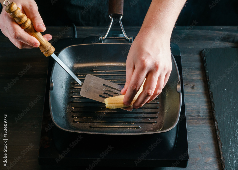Chef fries a peeled banana on an electric grill pan, helping himself with a kitchen spatula. The frying pan is placed on a black table top made of textured wood.