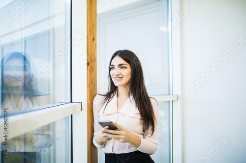 Young woman busy using her mobile phone while standing in front of large glass windows Free Download Young woman busy using her mobile phone while standing in front of large glass windows - GFXLoad