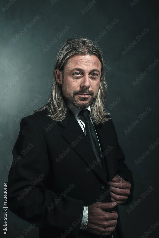 Retro victorian style man with blond long hair and beard. Wearing black suit and tie.