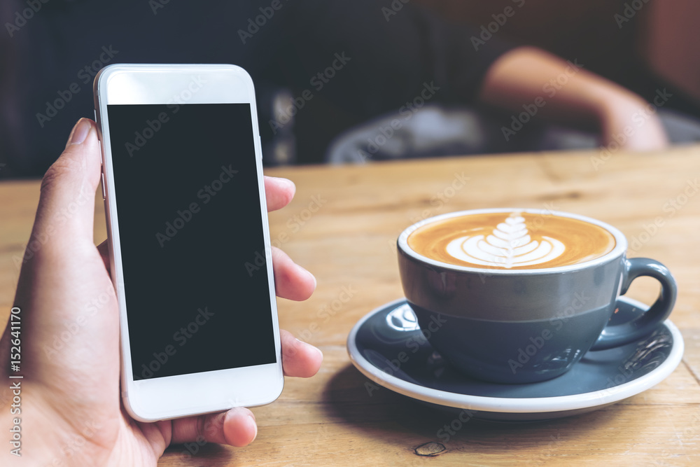 Mockup image of hand holding white mobile phone with blank black screen and hot coffee cup on vintage wooden table in cafe