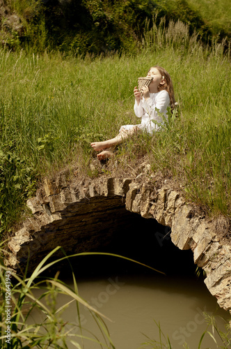 Portrait of little Caucasian girl in white dress seating on old little stone bridge and playing the flute (Palmanova, Friuli-Venezia Giulia Italy)