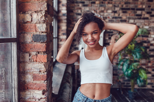 Beautiful african girl in white top standing near the window. © kanashkin