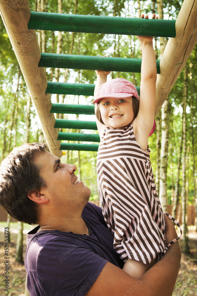 Obraz premium Father helping her daughter hanging on on the monkey bars in a playground, smiling and spending time together.