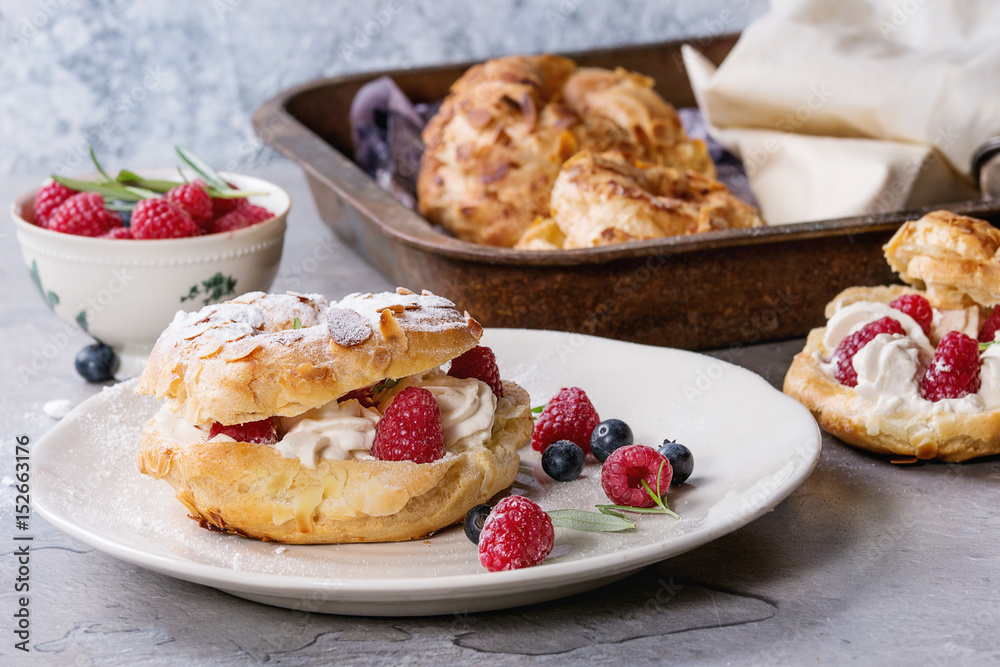 Filling and empty homemade choux pastry cake Paris Brest with raspberries, almond, sugar powder, rosemary on plate and oven tray with berries over gray texture background. French dessert. Filling and empty homemade choux pastry cake Paris Brest with raspberries, almond, sugar powder, rosemary on plate and oven tray with berries over gray texture background. French dessert.