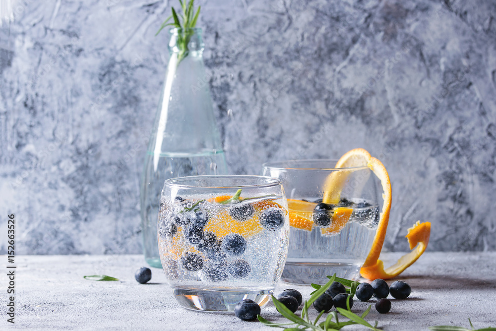 Tonic water cocktail with rosemary, blueberries and orange. Two cold glasses and bottle with zest and bubbles over gray blue texture background. Refreshing beverage alco non alcohol
