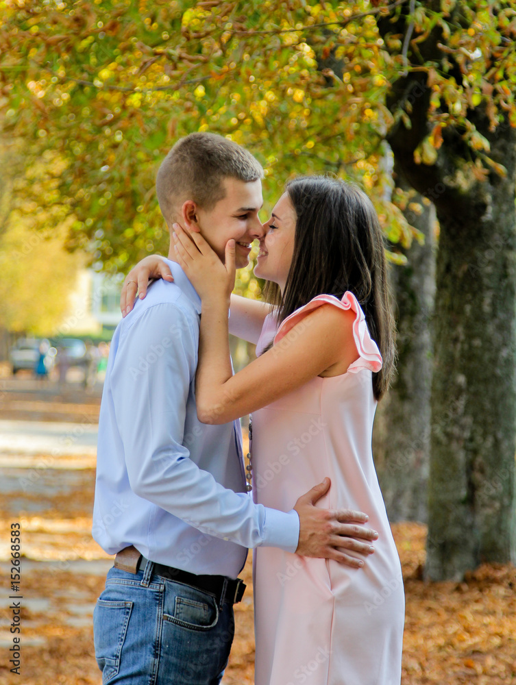 Happy Couple hugs and kissing. The warm and orange autumn and beautiful tree. Girl dressed in pink dress and man in blue shirt and jeans. Walking in the park Happy Couple hugs and kissing. The warm and orange autumn and beautiful tree. Girl dressed in pink dress and man in blue shirt and jeans. Walking in the park