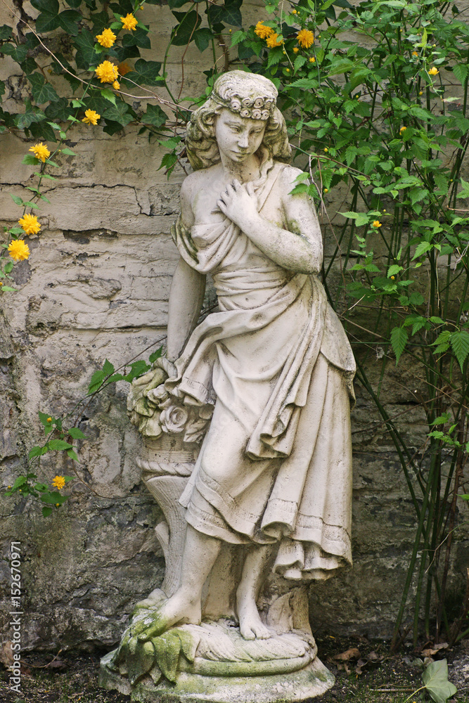 Statue eines Maedchens aus Stein auf einem Sockel vor einer Mauer