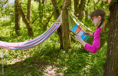 A girl in a hammock is reading a book in glasses.
