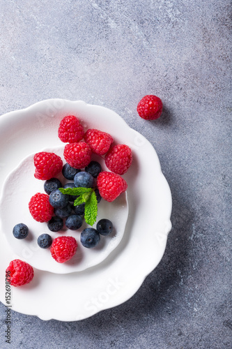 Freshly picked blueberries and raspberries on white plate on gray stone background. Concept for healthy eating and nutrition with copy space. Top view.