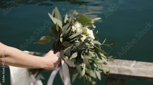 The hand is holding the bouquet consisted of the white field flowers and bay leaves placed on the balcony fence at the background of the blue river. photo