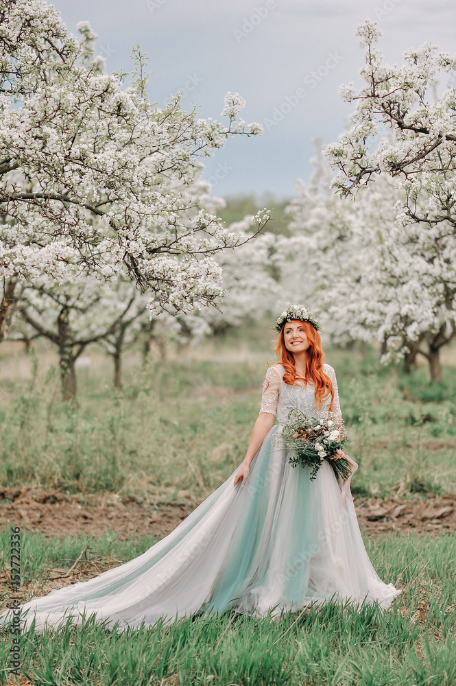 Young red-haired woman in a luxurious dress is standing in a blooming garden