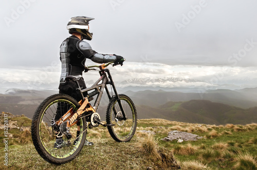 The rider in the full-face helmet and full protective equipment on the mtb bike stands on a rock against the background of a ridge and low clouds