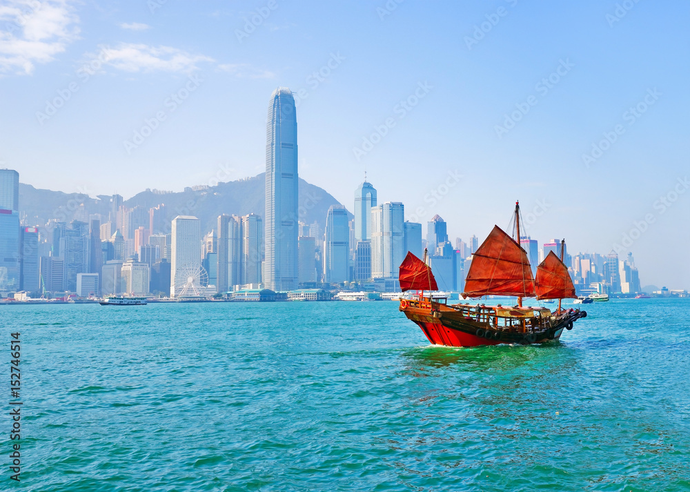 Obraz premium Widok na panoramę Hongkongu z czerwoną chińską żaglówką przechodzącą na Port Wiktorii w słoneczny dzień.