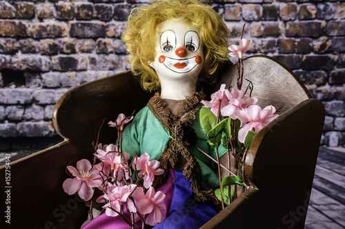 porcelain clown doll in antique wood cradle holding pink flowers with brick wall background