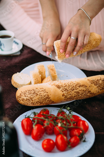 Girl cut baguette on a plate next to cherry tomatoes, white cup of coffee and a basket. photo