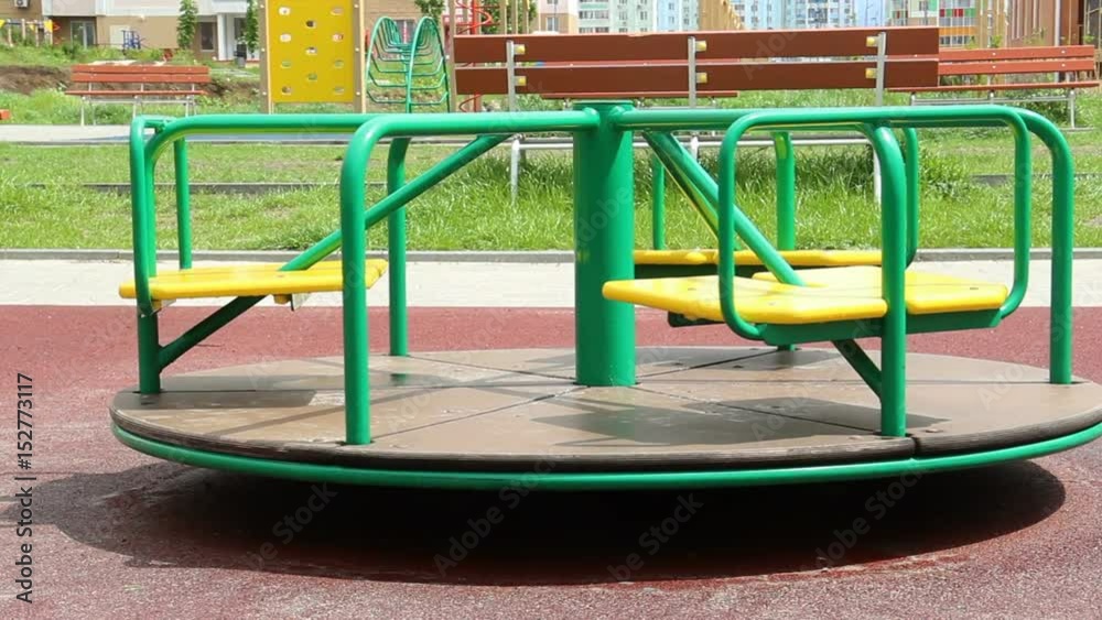 Yellow with green color, a wet carousel is spinning alone on a rubberized burgundy playground. no people. In the background high residential buildings of high-rise buildings Yellow with green color, a wet carousel is spinning alone on a rubberized burgundy playground. no people. In the background high residential buildings of high-rise buildings