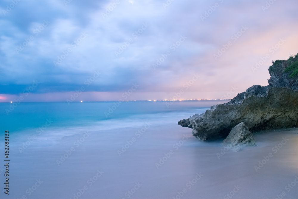 Long exposure photography of beach in the evening. Romantic atmosphere of peaceful night at sea, Bali island. Big rocks near smooth shoreline, pink horizon with sun rays, Indonesia outdoor landscape