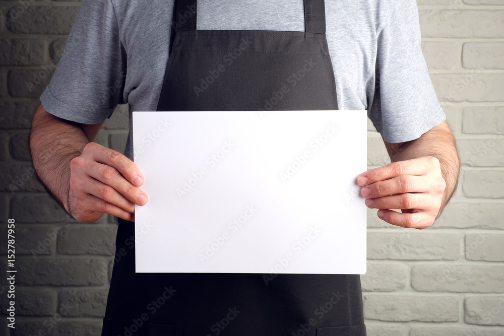 man in black apron holds white sheet of paper in his hands, on background of brick wall mock up man in black apron holds white sheet of paper in his hands, on background of brick wall mock up