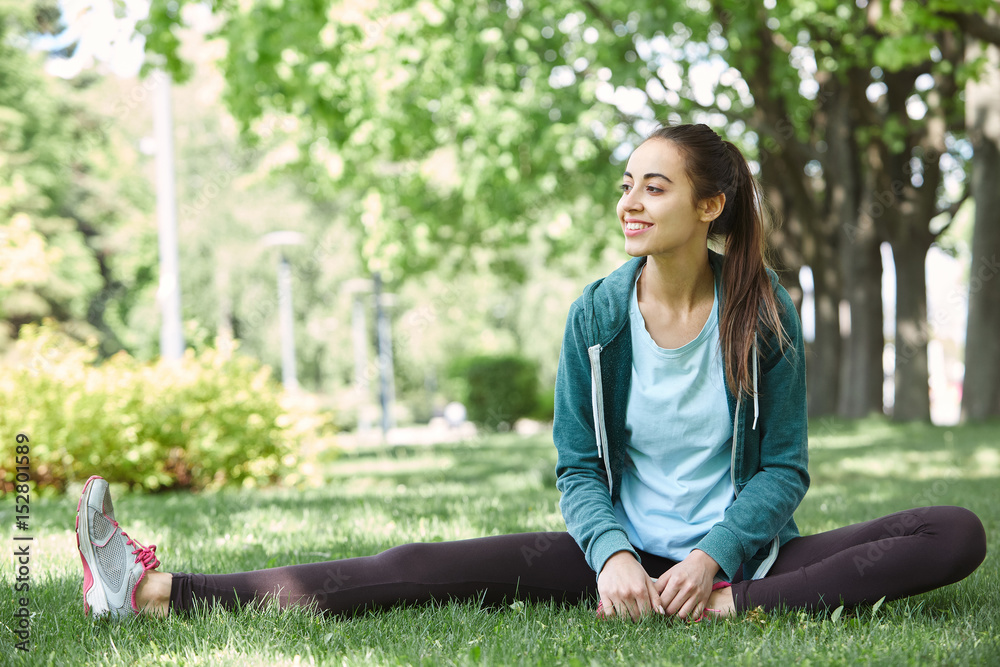 Portrait of young and sporty woman in sportswear doing yoga or Stretching exercises outside at the park on green field on cloudy day, Dnipro, Ukraine. She is sitting on the green meadow between trees Portrait of young and sporty woman in sportswear doing yoga or Stretching exercises outside at the park on green field on cloudy day, Dnipro, Ukraine. She is sitting on the green meadow between trees