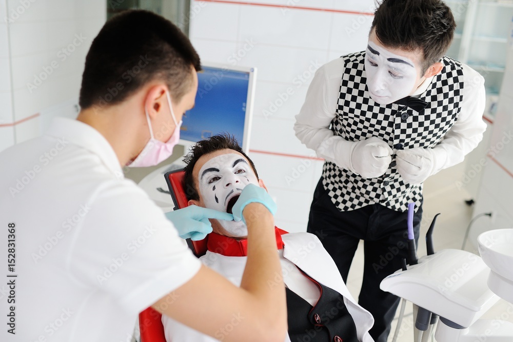 Fototapeta premium The clown or mime is sitting in the dental chair. The dentist treats the tooth to the patient.
