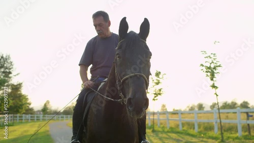 SLOW MOTION CLOSE UP, DOF: Portrait of an elder man horseback riding beautiful dark brown gelding at golden light sunset. Silhouetted senior rider mounted on a stallion standing still on a horse ranch