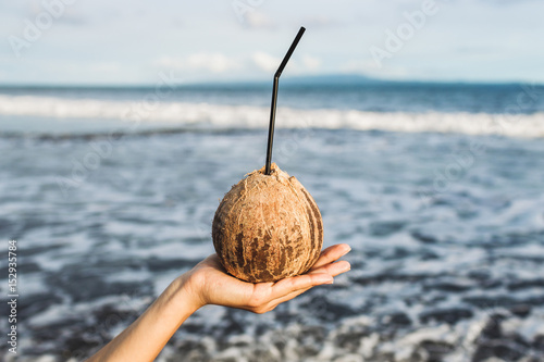 Woman's hand holding coconut at ocean coast in Bali. Blue tropical background with empty space for logo. Cold and healthy drink in summer