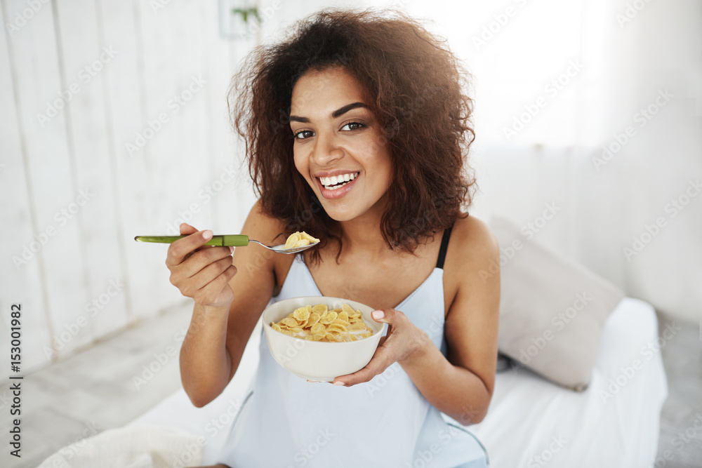 Beautiful african girl in sleepwear smiling looking at camera eating flakes with milk at home in the morning. Copy space.