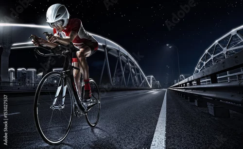 Fototapeta samoprzylepna Desery sportowe. Sportowa kobieta jeździć na rowerze drogowego rower w wieczór. Dramatyczny widok na miasto nocą.