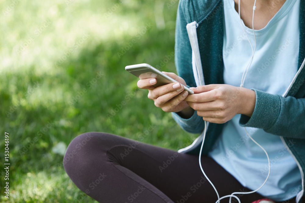 Portrait of young and sporty woman in sportswear sitting with the smartphone and headphones on the grass outside at the park on green field on cloudy day, Dnipro, Ukraine. She is sitting on the green