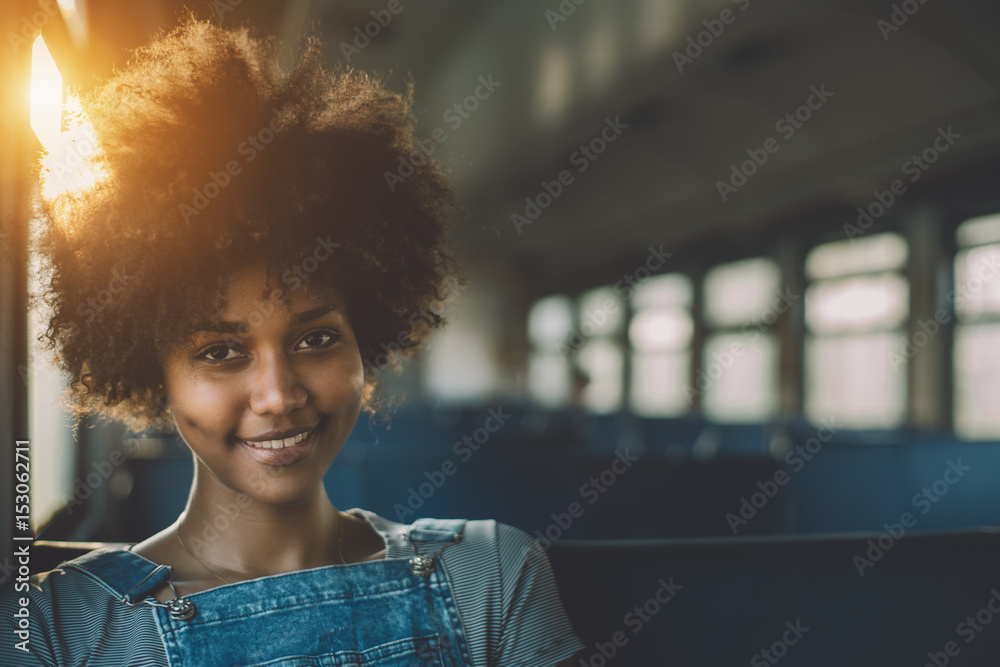 Young smiling beautiful black girl with curly afro hair sitting goes by suburban train, attractive mixed teenage female on seat of empty electrical train with area for your text message or logo