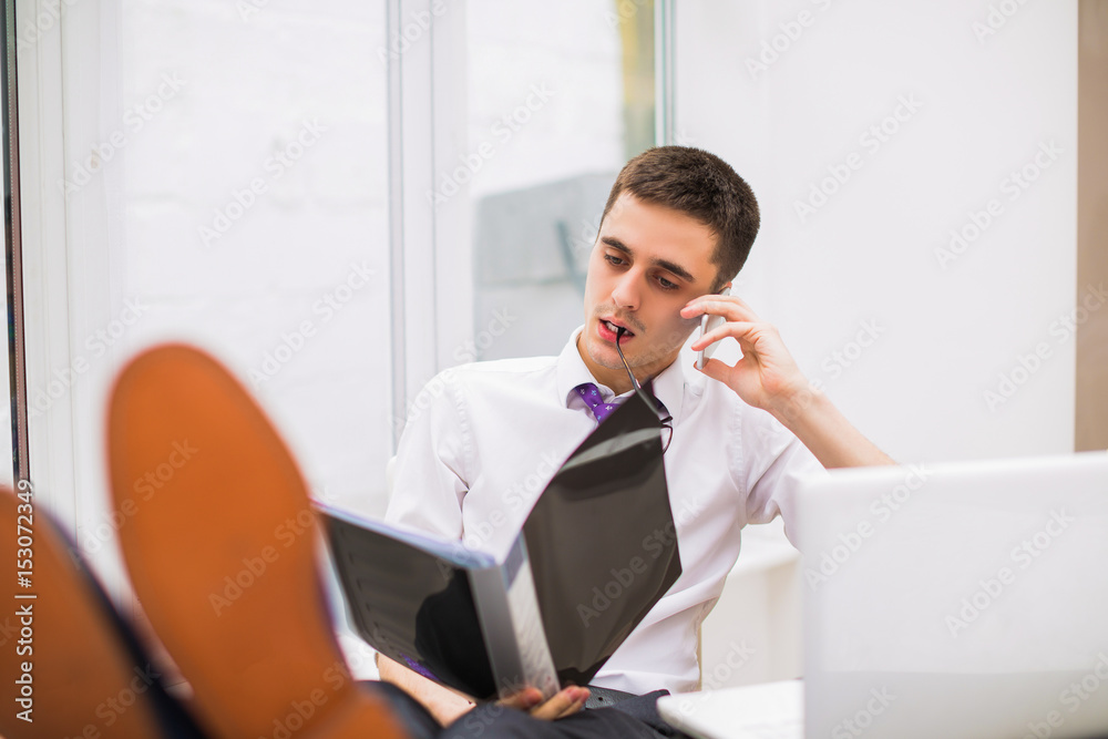 Young handsome manager sits, speaks the phone and looks at opened folder at the office Young handsome manager sits, speaks the phone and looks at opened folder at the office