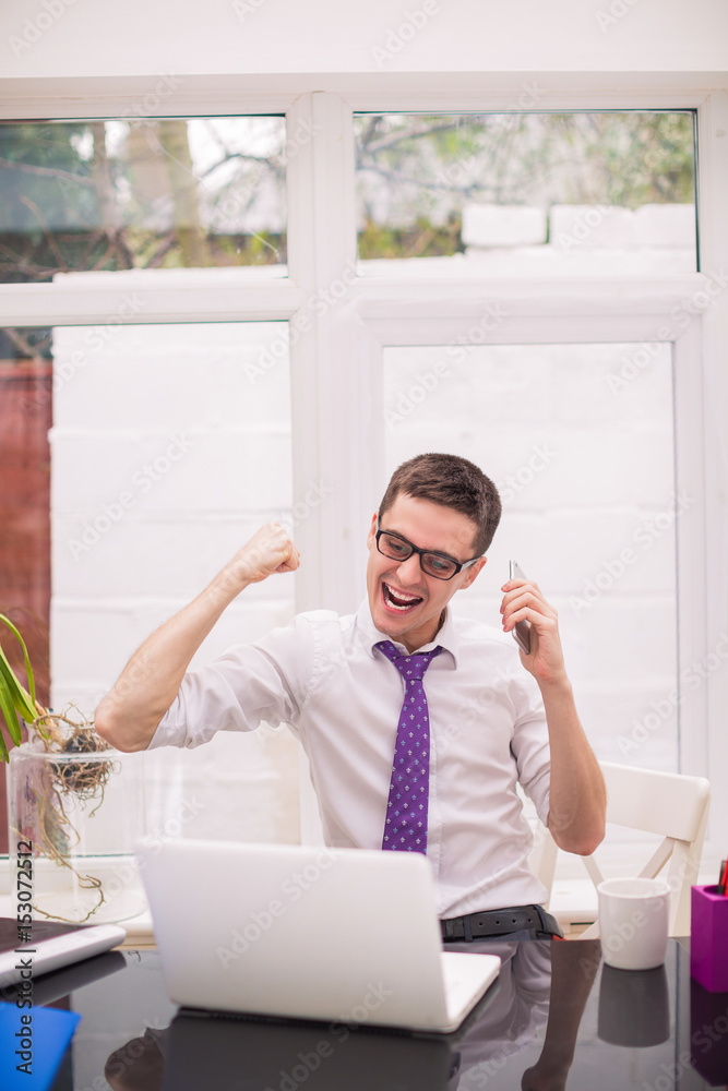 Young happy businessman looks at laptop, rejoices for something or someone and keeps the phone in hand at the office