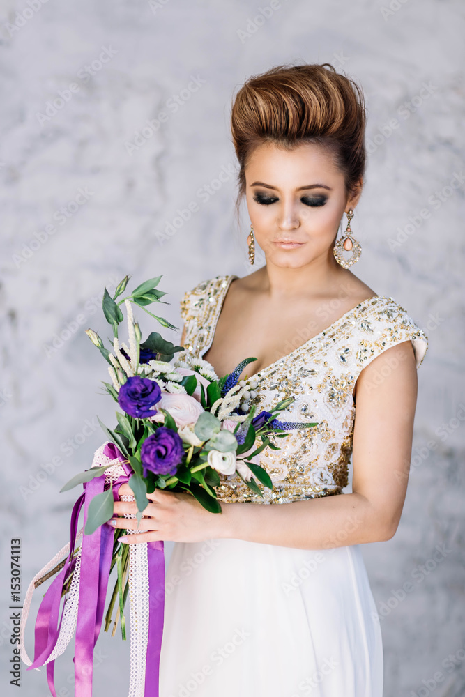 beautiful girl bride blond girl in white dress with gold decorations with wedding bouquet makeup and hair in the Studio looking down with a smile close-up white background beautiful girl bride blond girl in white dress with gold decorations with wedding bouquet makeup and hair in the Studio looking down with a smile close-up white background
