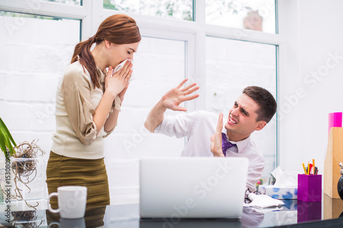 Young sick office worker stands near her colleague at the office and wipes nose