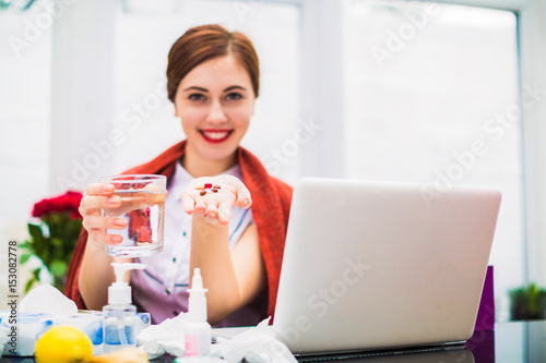 Portrait of sick business lady who sits at the working table, keeps several pills and cup of water at the office