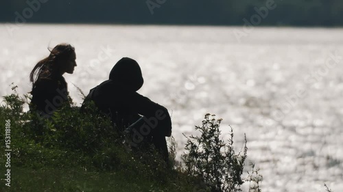 Couple silhouette talks siting on grass in front of water of a lake on a summer windy day. Man wearing hood, womans hairs waves in wind