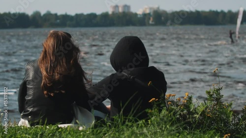 Couple sitting on grass watch as windsurfer rides across water in front of them on a summer windy day
