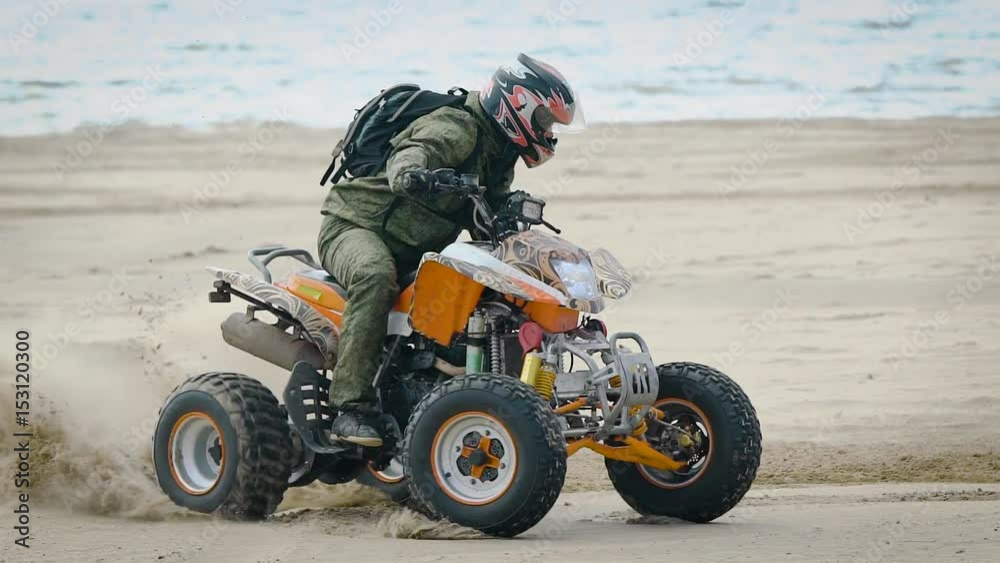 Close up of man in protective equipment and helmet drifting on his ATV on the beach with the sea in the background. Extreme athlete riding his quad bike and moving around dusting on the shore. Close up of man in protective equipment and helmet drifting on his ATV on the beach with the sea in the background. Extreme athlete riding his quad bike and moving around dusting on the shore.