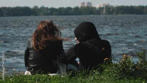 Couple silhouette talks siting on grass in front of water of a lake on a summer windy day. Man wearing hood, womans hairs waves in wind. City buildings across the shore.
