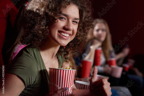 Side view of beautiful smiling woman with volumed curly hair looking, posing at camera at cinema hall, holding tasty popcorn. Young emotionally brunette girl watching funny movie at modern cinema.