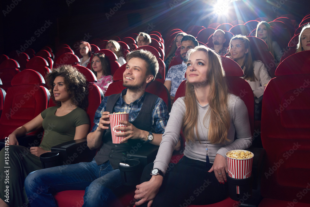 Smiling positive couple of boyfriend and blonde girlfriend watching funny movie at cinema . Best friends spending time together, eating popcorn when looking at project. Big fiends group.