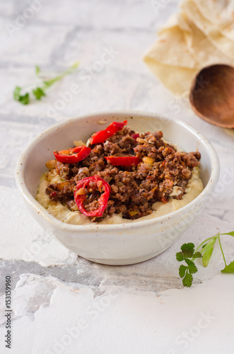Creamy hummus with spicy mutton minced meat in clay bowl on stone background. Ramadan food.
