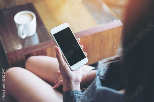 Mockup image of a woman holding white mobile phone with blank black screen with coffee mug on wooden table in cafe