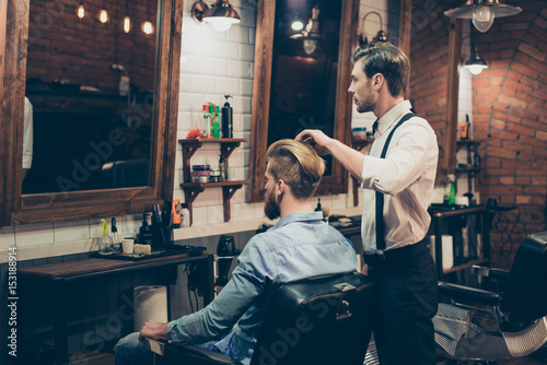 Barber shop classy dressed stylist is working for a perfect hairdo of a blond bearded guy in caual jeans outfit. Both looking in the mirror, concentrated