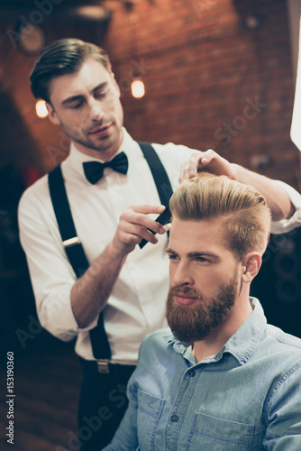 Barber shop classy dressed handsome stylist is doing a perfect hairstyle to a bearded guy in caual jeans outfit. Both concentrated and serious