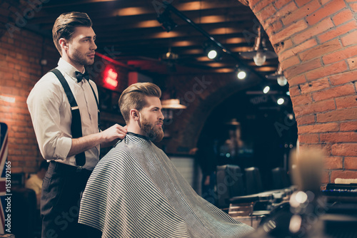 Great job! Hairstylist and a client in a barber shop. Red bearded young man got a stunning hairdo and his beard got cutted perfectly. Hairdresser is taking off the cape