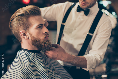 Cropped photo of a classy dressed barber shop stylist working for a perfect look of a red bearded guy in a cape. His hairdo looks stunning!