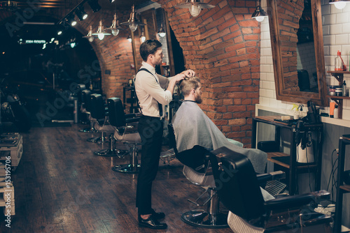 Barber shop classy dressed specialist is styling the hair of a client. Salon is retro and vintage. Customer is a young bearded man, covered with cape