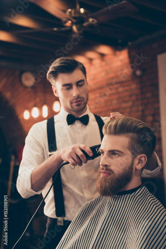 Beard styling for a handsome young guy at the barber shop. Hairdresser is attractive and wearing classic outfit, he is focused and serious