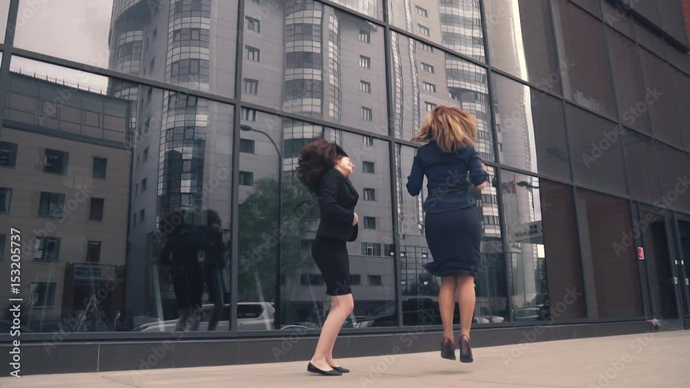 two girls in the clothing business is happy, jumping and having fun. business woman celebrating success and victory. businessmen on background of office building of glass. slow motion two girls in the clothing business is happy, jumping and having fun. business woman celebrating success and victory. businessmen on background of office building of glass. slow motion